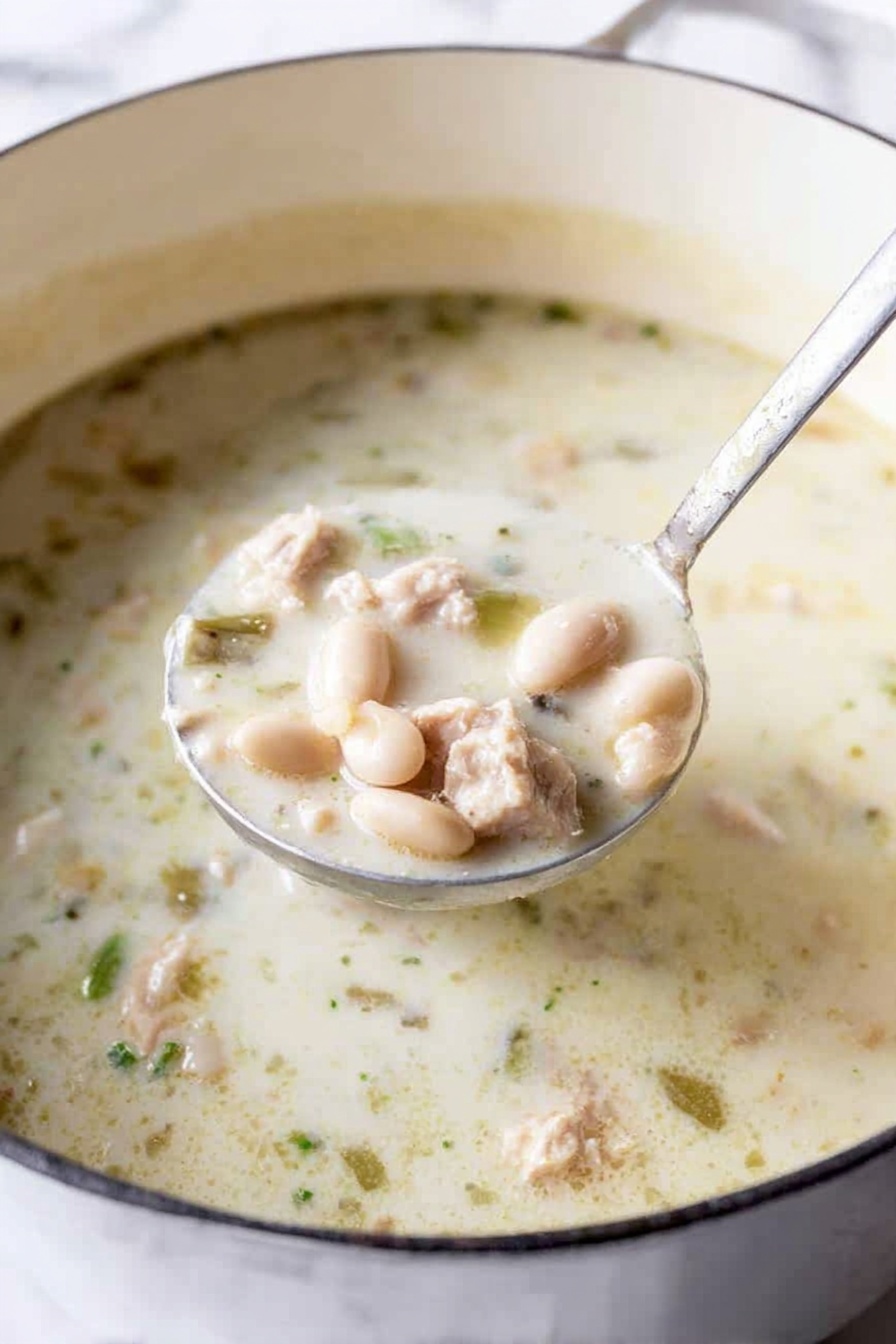 Creamy White Chicken Chili, White Chicken Chili recipe, easy chicken chili, comforting chicken chili, homemade white chili - The image shows a large white pot filled with creamy white soup that has a slightly thick texture. The soup contains small white beans, light beige pieces of tender meat, and small green vegetable bits, all mixed evenly throughout the creamy broth. A metal ladle is lifting a portion of the soup, displaying a close-up of the ingredients which include the beans, meat, and green bits clearly visible in the rich white soup. The pot’s rim is white with a thin dark edge, and the background is a white marbled texture. photo taken with an iphone --ar 2:3 --v 7