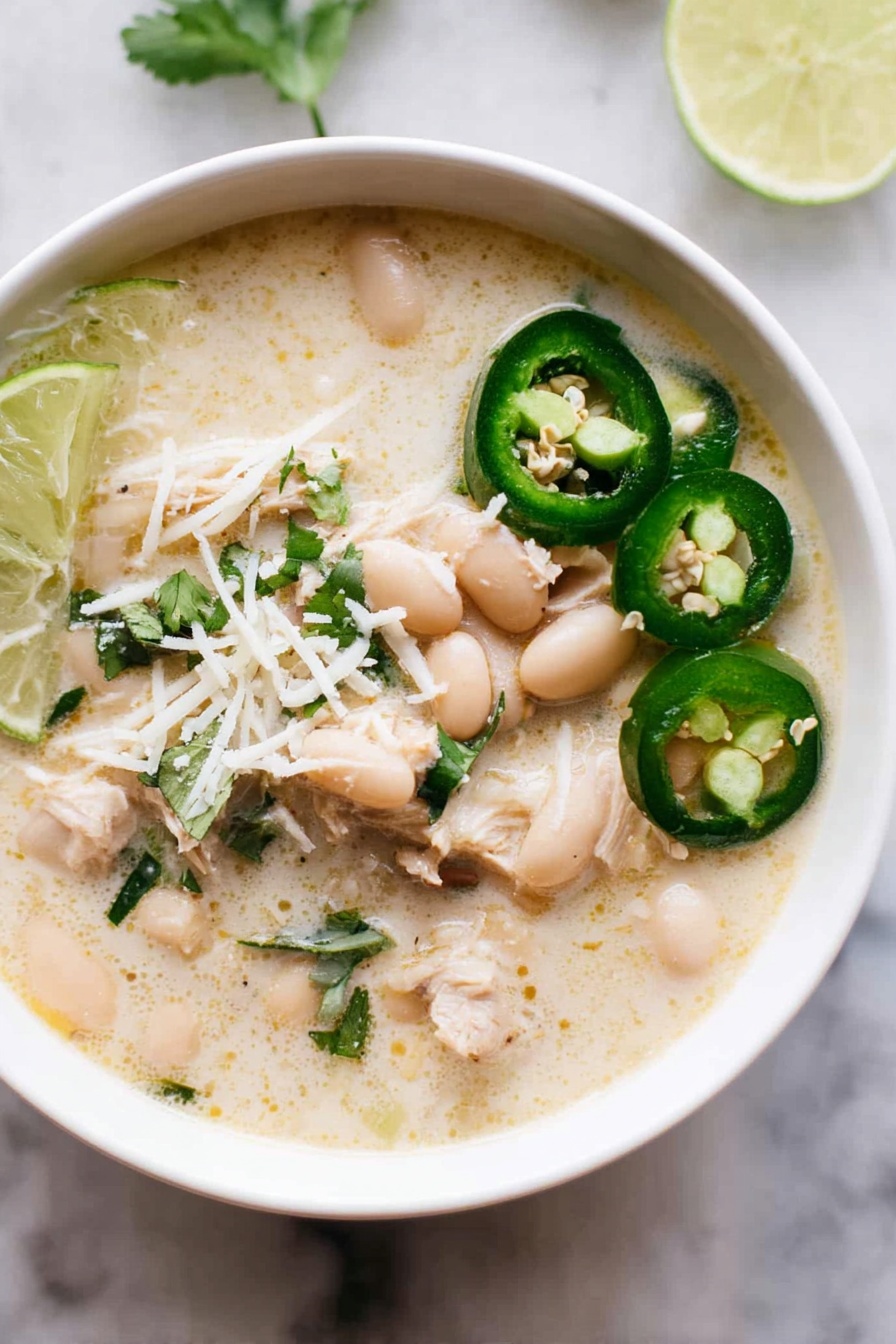Creamy White Chicken Chili, White Chicken Chili recipe, easy chicken chili, comforting chicken chili, homemade white chili - A close-up view of a creamy white soup served in a white bowl, showing three main layers: a broth layer that is thick and pale cream in color with specks of green herbs; a layer of soft, shredded light beige chicken pieces scattered throughout the soup; and a layer of smooth, oval white beans floating on top. On the surface, there are thin slices of bright green jalapeño peppers placed near the edge of the bowl, along with a wedge of fresh lime with a vibrant green rind. Some chopped green herbs are sprinkled across the soup for color. A shiny metal spoon scoops a portion of the soup, holding a mix of chicken, beans, and broth. The bowl is set on a white marbled surface. Photo taken with an iphone --ar 2:3 --v 7
