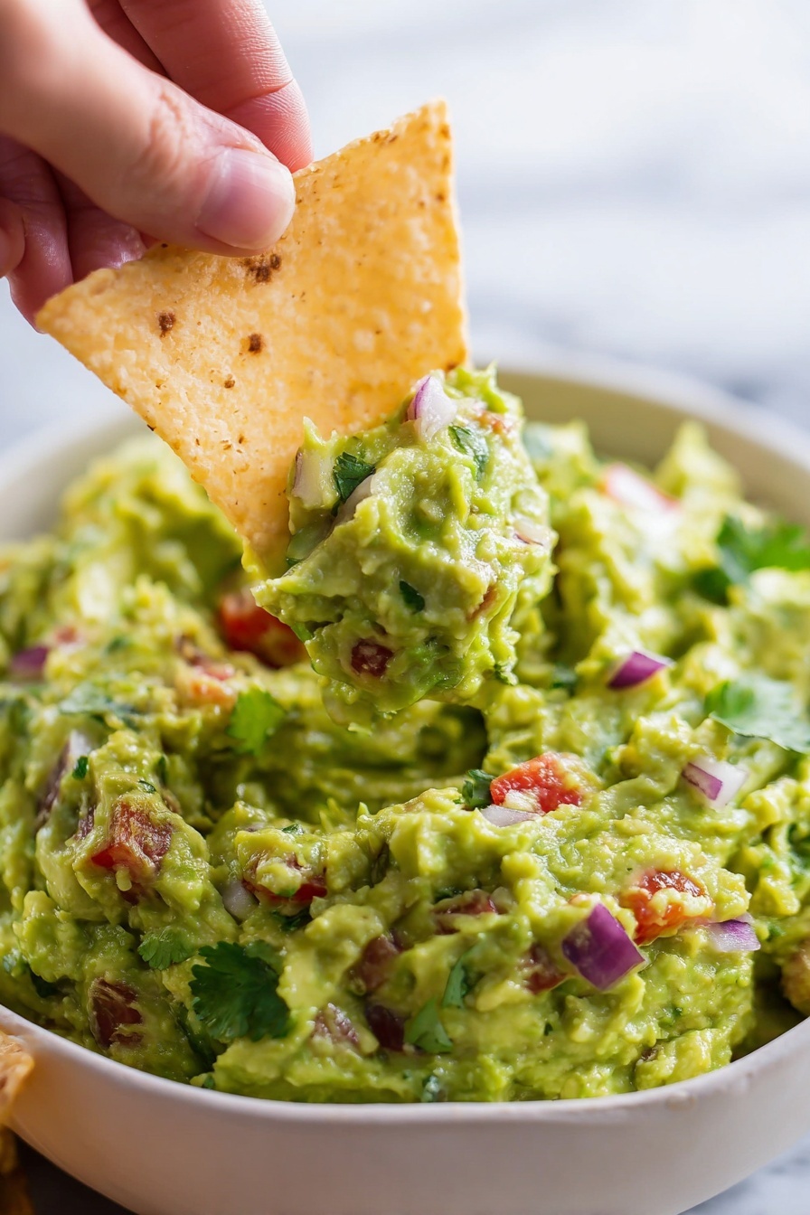 Homemade Guacamole with Fresh Avocados, how to make fresh guacamole, easy guacamole recipe, quick avocado dip, healthy homemade guacamole - The image shows a speckled white bowl filled with freshly made guacamole that has a chunky texture. The guacamole is bright green with visible small pieces of red tomatoes, purple onions, and green cilantro mixed throughout. Three crispy, triangular tortilla chips are partially dipped vertically into the guacamole on one side of the bowl. More tortilla chips are scattered around the bowl on a white marbled surface, adding a crunchy contrast to the creamy dip. photo taken with an iphone --ar 2:3 --v 7