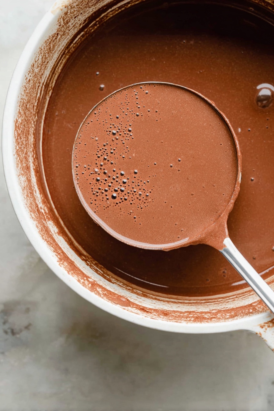 Mexican Hot Chocolate, authentic Mexican hot chocolate, spicy hot chocolate recipe, easy Mexican hot chocolate, rich cinnamon hot chocolate - A close-up view of a ladle filled with smooth, light brown liquid chocolate mixture held above a white pot containing the same liquid. The mixture has a creamy texture with tiny bubbles and slight froth on top. The edge of the pot shows splashes of the chocolate mixture, and the background surface is a white marbled texture. photo taken with an iphone --ar 2:3 --v 7