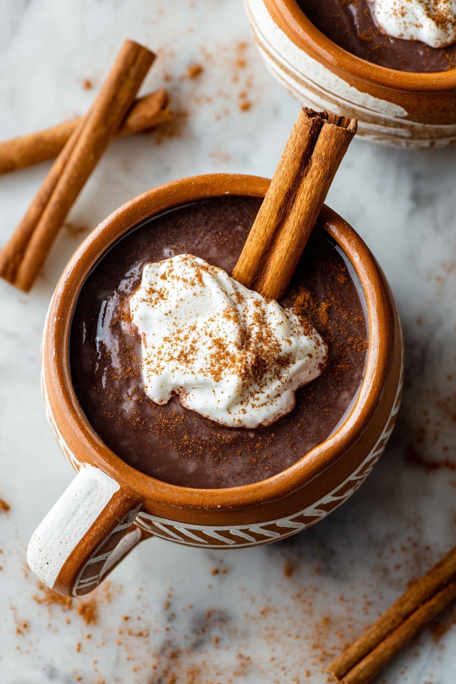 Mexican Hot Chocolate, authentic Mexican hot chocolate, spicy hot chocolate recipe, easy Mexican hot chocolate, rich cinnamon hot chocolate - The image shows two round clay mugs filled with dark brown hot chocolate. Each mug has a dollop of white whipped cream on top, sprinkled with light brown cinnamon powder. Two long cinnamon sticks are placed upright inside the mug in the front, leaning against the whipped cream. The mugs have white painted lines on the outside. The mugs are placed on a white marbled surface with some scattered cinnamon sticks and cinnamon powder around them. The shot is close up, focusing on the mug in the front, with soft natural light highlighting the textures. Photo taken with an iphone --ar 2:3 --v 7