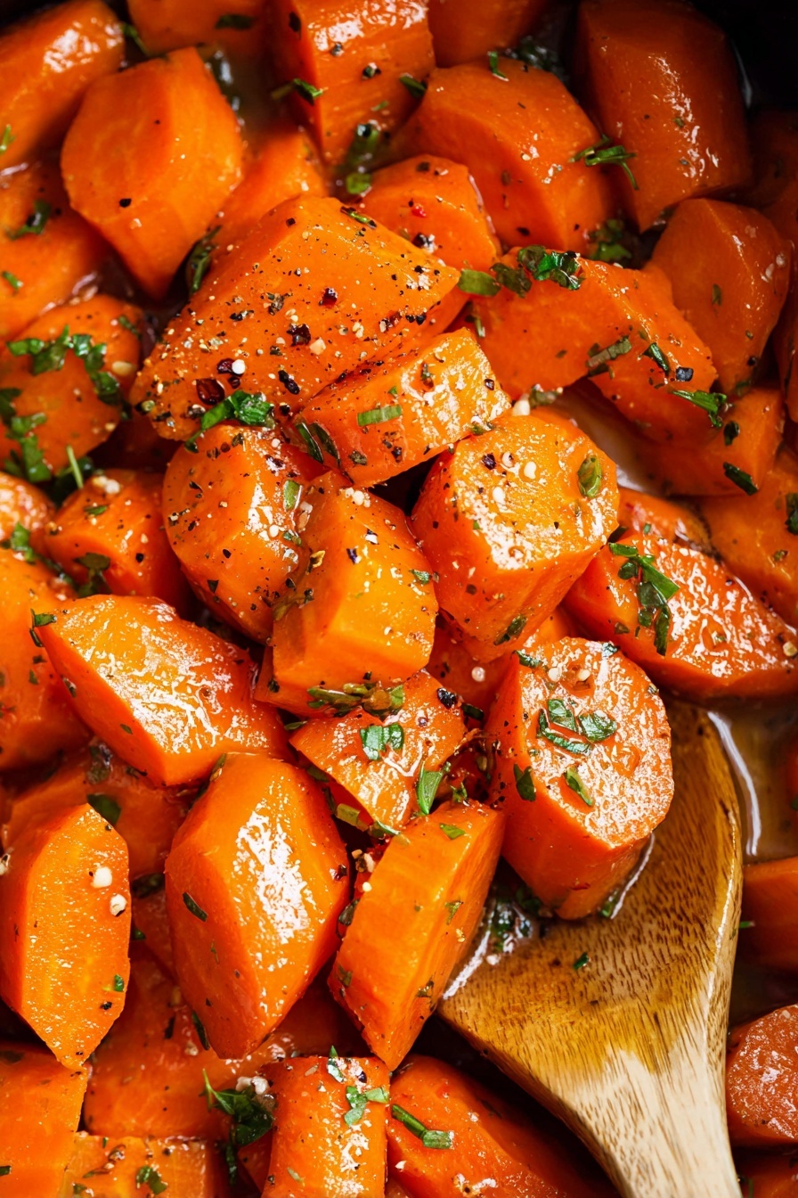 Sweet Honey Garlic Crockpot Carrots, honey garlic carrots, slow cooker carrot side dish, easy healthy carrot recipes, tender glazed carrots - A close-up image showing a bowl filled with bright orange cooked carrot pieces. The carrots are cut into irregular chunks with some pieces rounded and others more angular. They have a shiny, slightly glazed texture with small specks of black pepper scattered on top along with finely chopped green parsley. A gold spoon rests in the bowl, partially covered by carrots. The bowl is white with a thin brown rim and is placed on a white marbled surface with a few green herb leaves nearby. Photo taken with an iphone --ar 2:3 --v 7